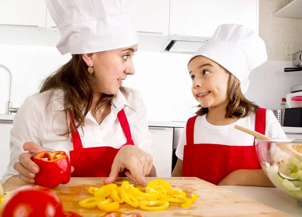 Happy mother and little daughter at home kitchen preparing paprika salad in apron and cook hat — Stockfoto