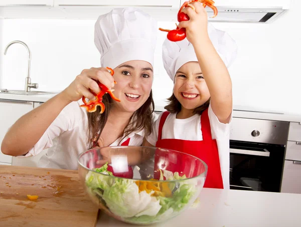 Happy mother and little daughter at home kitchen preparing paprika salad in apron and cook hat — Stockfoto