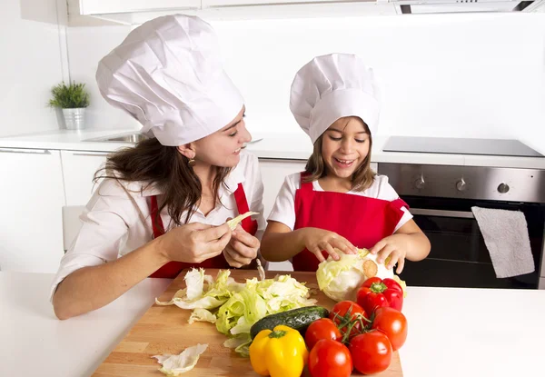 Happy mother and little daughter at home kitchen preparing salad in apron and cook hat — Stok fotoğraf