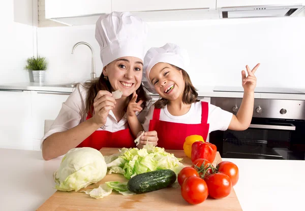 Feliz madre e hija pequeña en la cocina casera preparando ensalada en delantal y sombrero de cocinero —  Fotos de Stock
