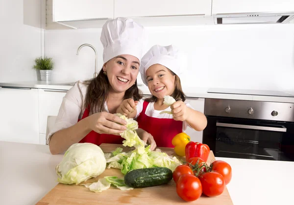 Happy mother and little daughter at home kitchen preparing salad in apron and cook hat — Zdjęcie stockowe