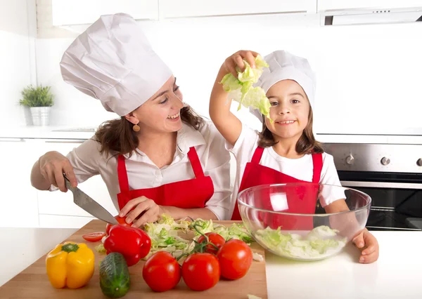 Young mother and little daughter at house kitchen preparing salad for lunch wearing apron and cook hat — Stockfoto