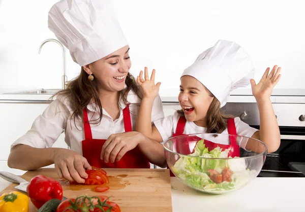 Young mother and little daughter at house kitchen preparing salad for lunch wearing apron and cook hat — Stock Photo, Image