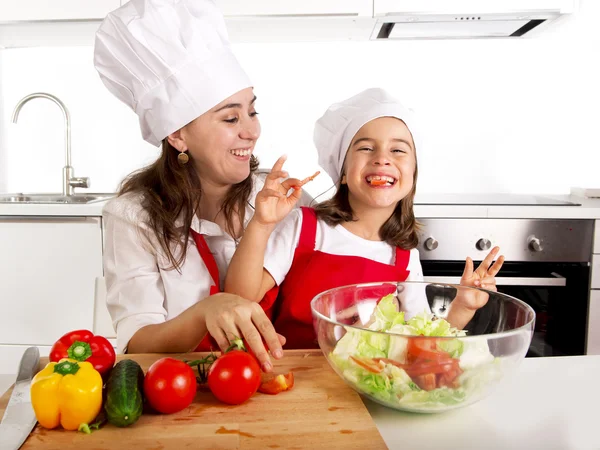 Young mother and little daughter at house kitchen preparing salad for lunch wearing apron and cook hat — Stock Photo, Image