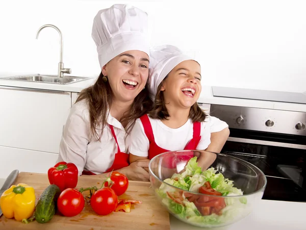 Young mother and little daughter at house kitchen preparing salad for lunch wearing apron and cook hat — Stockfoto