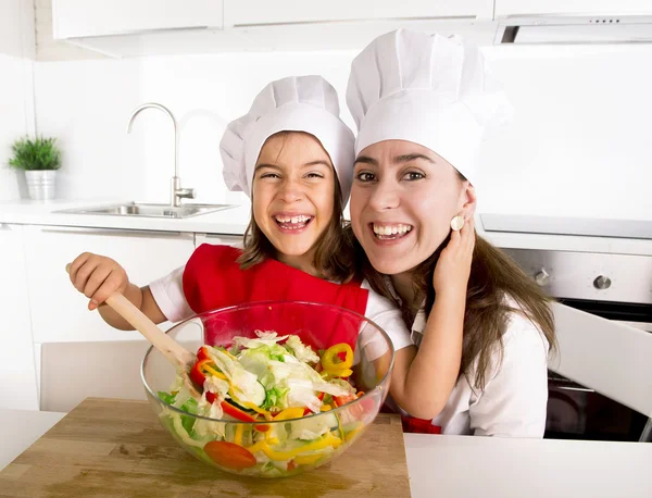 Mãe feliz e filha pequena em casa cozinha preparando salada em avental e cozinhe chapéu — Fotografia de Stock