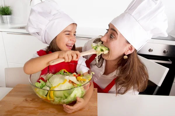 Happy mother and little daughter at home kitchen preparing salad in apron and cook hat — Stockfoto