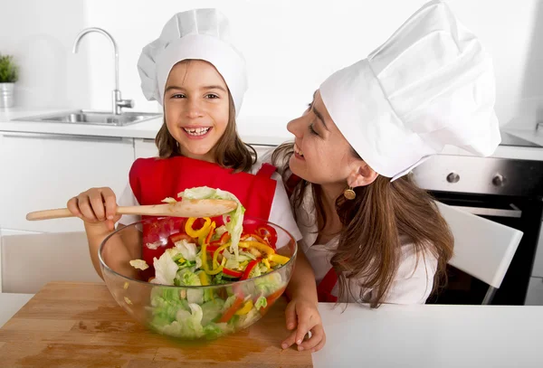 Happy mother and little daughter at home kitchen preparing salad in apron and cook hat — Stock Photo, Image