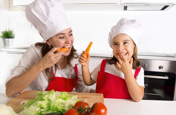 Happy mother and little daughter in apron and cook hat eating carrots together having fun at home kitchen — Zdjęcie stockowe