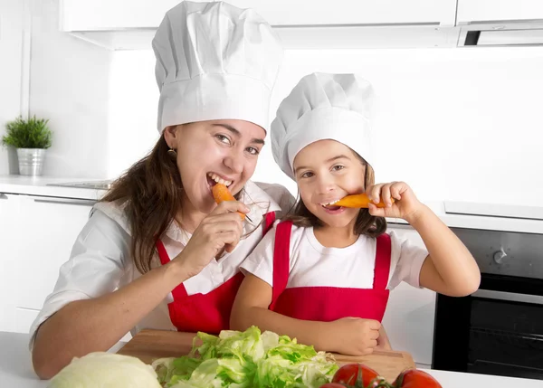 Happy mother and little daughter in apron and cook hat eating carrots together having fun at home kitchen — Stock Photo, Image