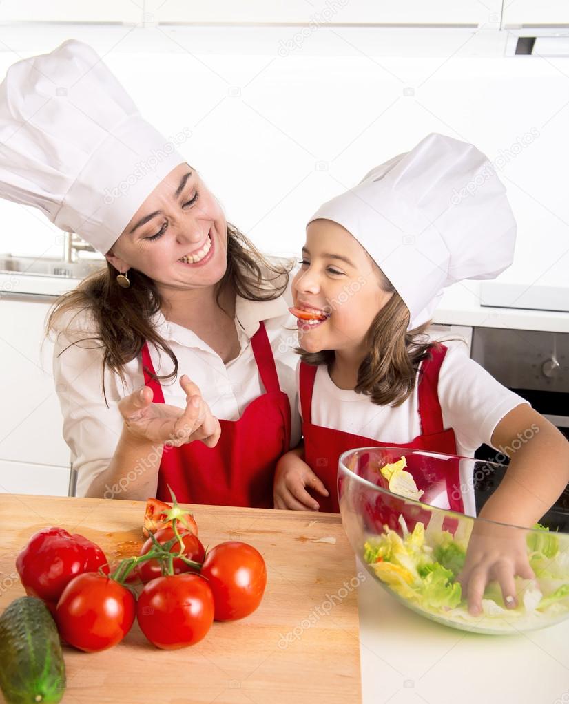 young mother and little daughter at house kitchen preparing salad for lunch wearing apron and cook hat