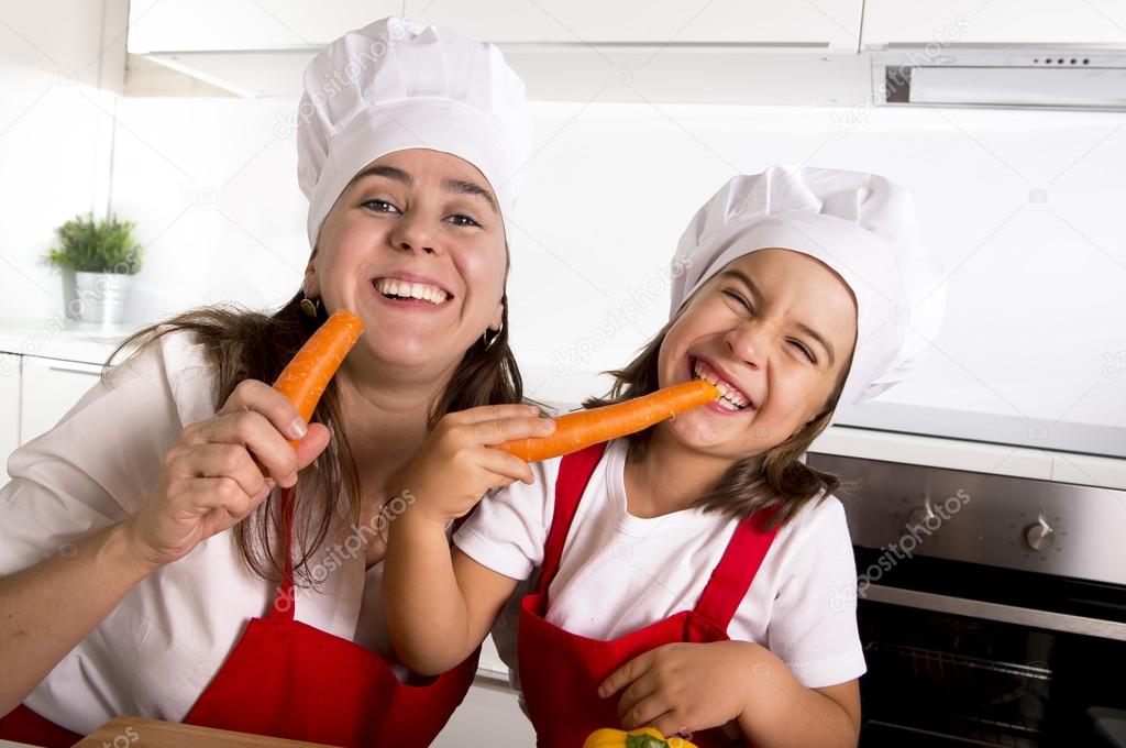 happy mother and little daughter in apron and cook hat eating carrots together having fun at home kitchen