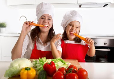 happy mother and little daughter in apron and cook hat eating carrots together having fun at home kitchen
