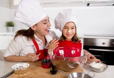 happy mother baking with little daughter in apron and cook hat with flour dough at kitchen