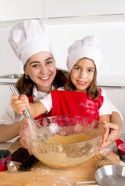 happy mother baking with little daughter in apron and cook hat preparing dough at kitchen