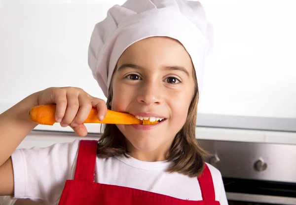 Gadis kecil di dapur rumah dengan celemek merah dan topi masak memegang wortel dan menggigit bahagia — Stok Foto