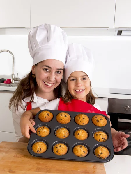Happy mother with daughter wearing apron and cook hat presenting muffin set baking together at home kitchen — Stock Photo, Image