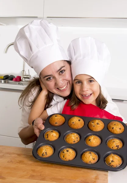 Happy mother with daughter wearing apron and cook hat presenting muffin set baking together at home kitchen — Stock Photo, Image