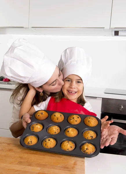 Happy mother with daughter wearing apron and cook hat presenting muffin set baking together at home kitchen — Stock Photo, Image