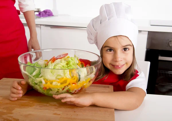 Sweet little girl  at home kitchen in red apron and cook hat holding vegetable salad bowl — Stock Photo, Image