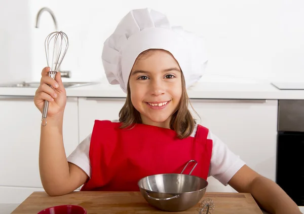 Sweet little girl in red apron and cook hat playing chef learning cooking at home kitchen — Stock Photo, Image