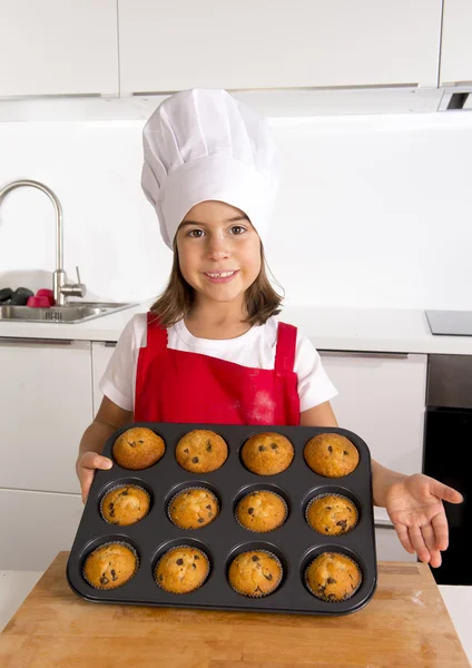 Proud female child presenting her self made muffin cakes learning baking wearing red apron and cook hat smiling happy — Stock Photo, Image