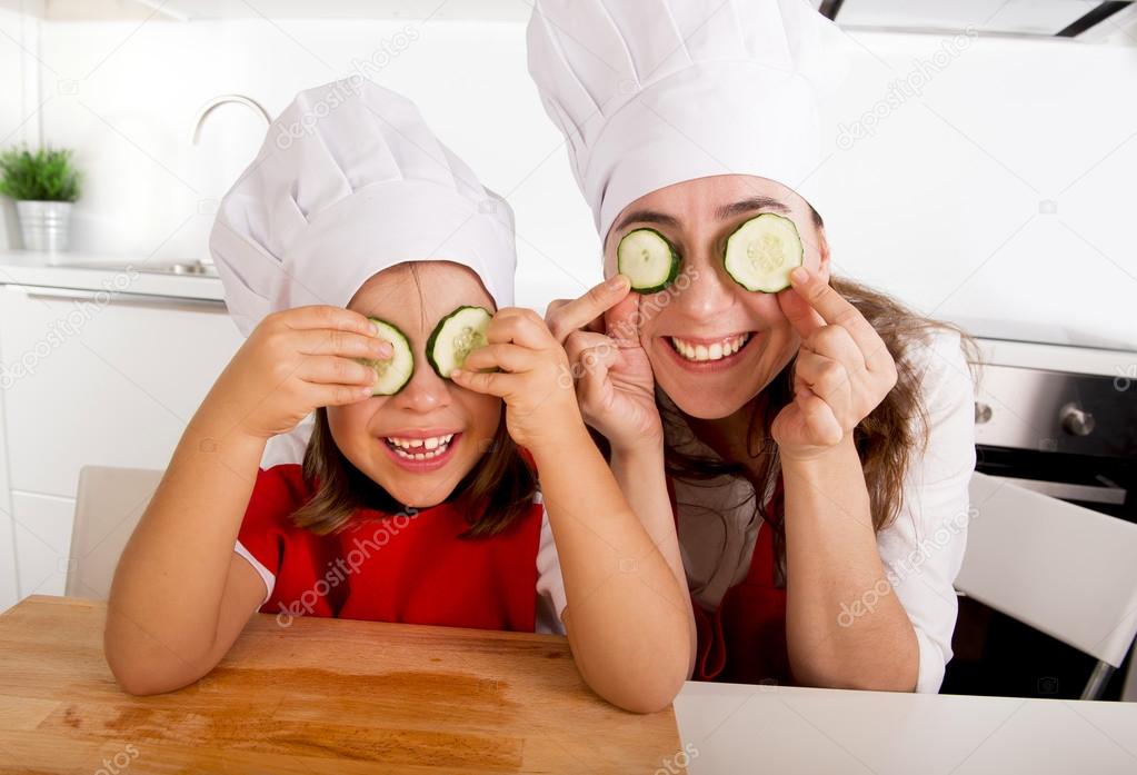 mother and little daughter in cook hat and apron playing with cucumber slices on eyes in kitchen