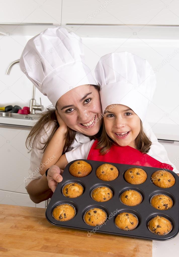 happy mother with daughter wearing apron and cook hat presenting muffin set baking together at home kitchen