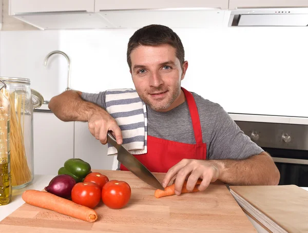 Junger Hausmann in Schürze schneidet Möhre mit Küchenmesser glücklich und entspannt — Stockfoto