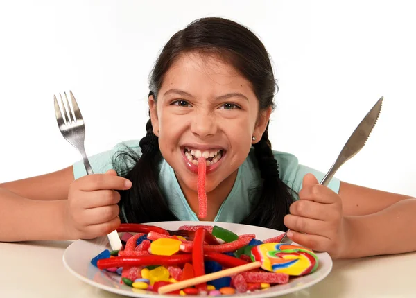 Ευτυχισμένος ενθουσιασμένος λατινικό θηλυκό παιδί κρατώντας πηρούνι και μαχαίρι που κάθεται στο τραπέζι είναι έτοιμο για να φάει ένα πιάτο γεμάτο από καραμέλα — Φωτογραφία Αρχείου