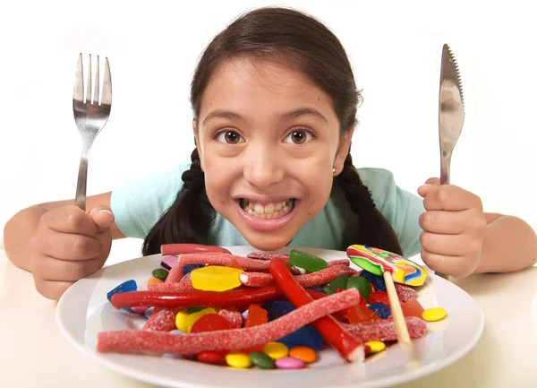 Boldog, izgatott, Latin női gyermek holding, Villa és kés, kész asztalnál eszik egy tál tele candy — Stock Fotó