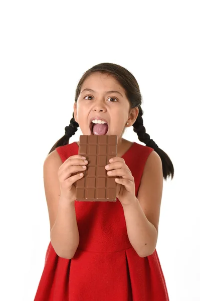 Hispanic female girl wearing red dress holding with both hands big chocolate eating in happy excited face expression in sugary nutrition — стокове фото