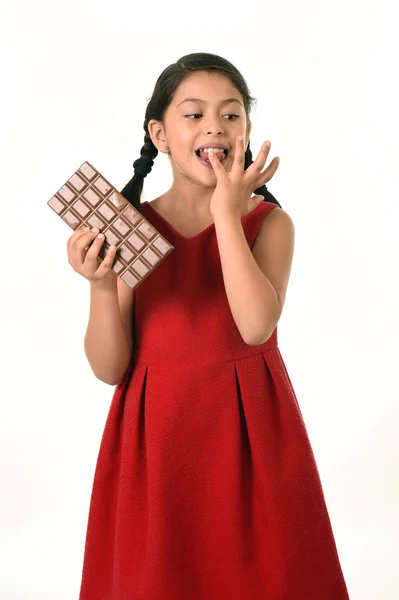 Hispanic female girl wearing red dress holding big chocolate bar eating in happy excited face expression licking her finger — Stok fotoğraf