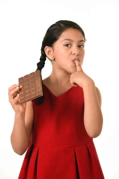 Hispanic female girl wearing red dress holding big chocolate bar eating in happy excited face expression licking her finger — Zdjęcie stockowe