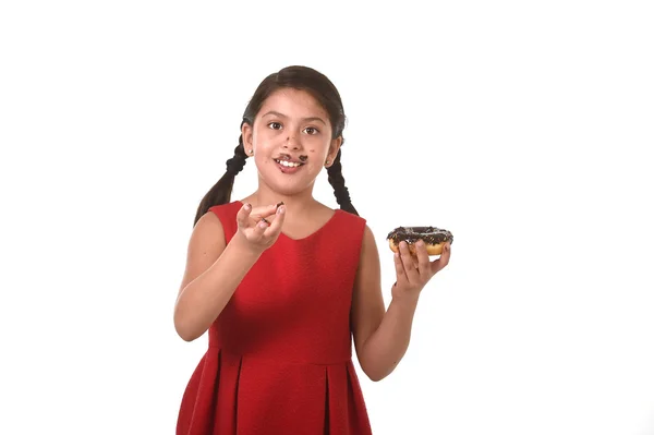 Niña latina en vestido rojo comiendo donut de chocolate con las manos y la boca manchada y sucia sonriente feliz — Foto de Stock