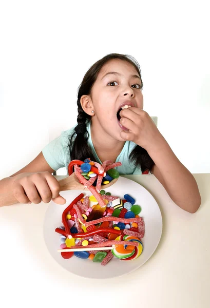 Felice ragazza che tiene cucchiaio mangiare da piatto pieno di caramelle lecca-lecca e cose zuccherine Immagine Stock