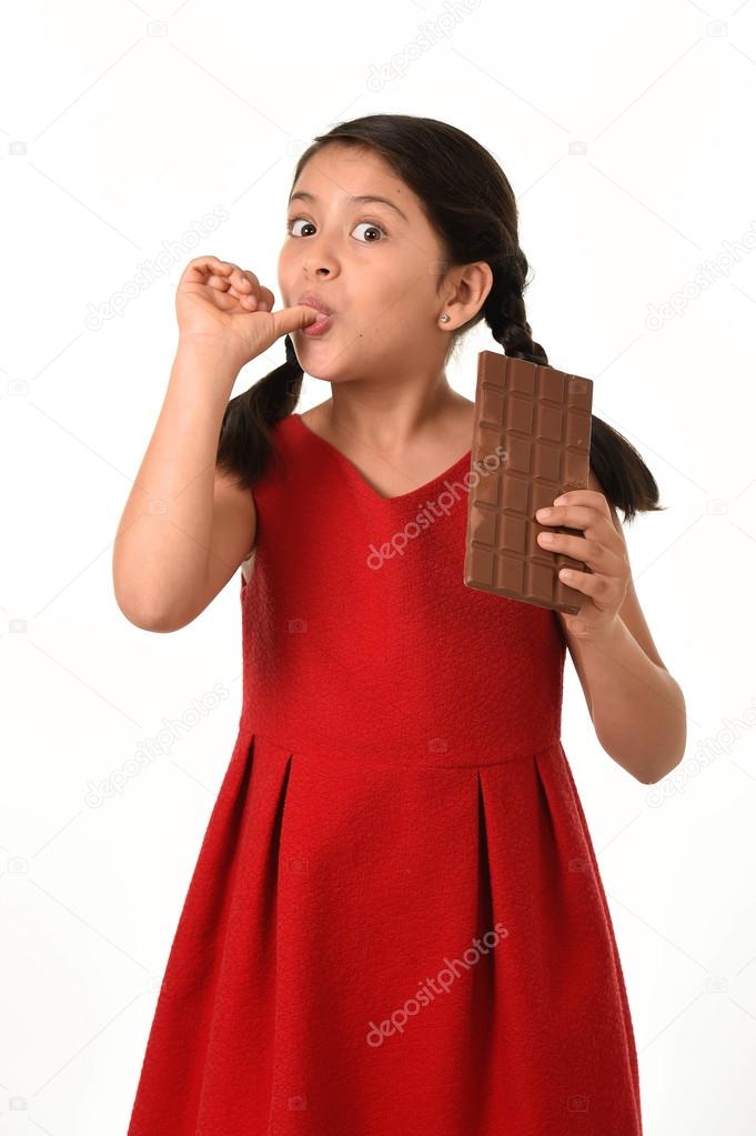 Hispanic female girl wearing red dress holding big chocolate bar eating in happy excited face expression licking her finger