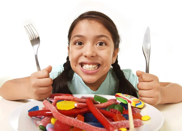 Feliz emocionado latina niña sosteniendo tenedor y cuchillo sentado a la mesa listo para comer un plato lleno de dulces —  Fotos de Stock