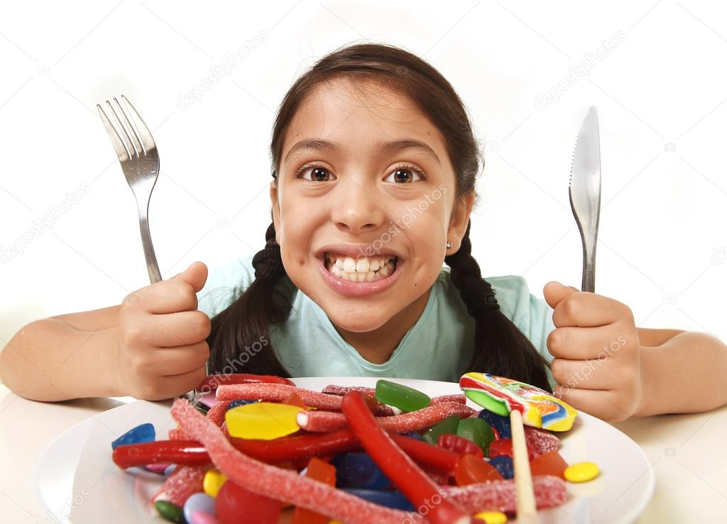happy excited Latin female child holding fork and knife sitting at table ready for eat a dish full of candy