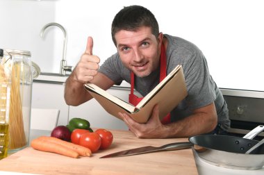 young happy man at kitchen reading recipe book in apron learning clipart