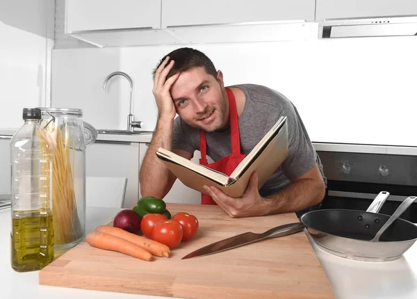 Young happy man at kitchen reading recipe book in apron learning — Stock Photo, Image