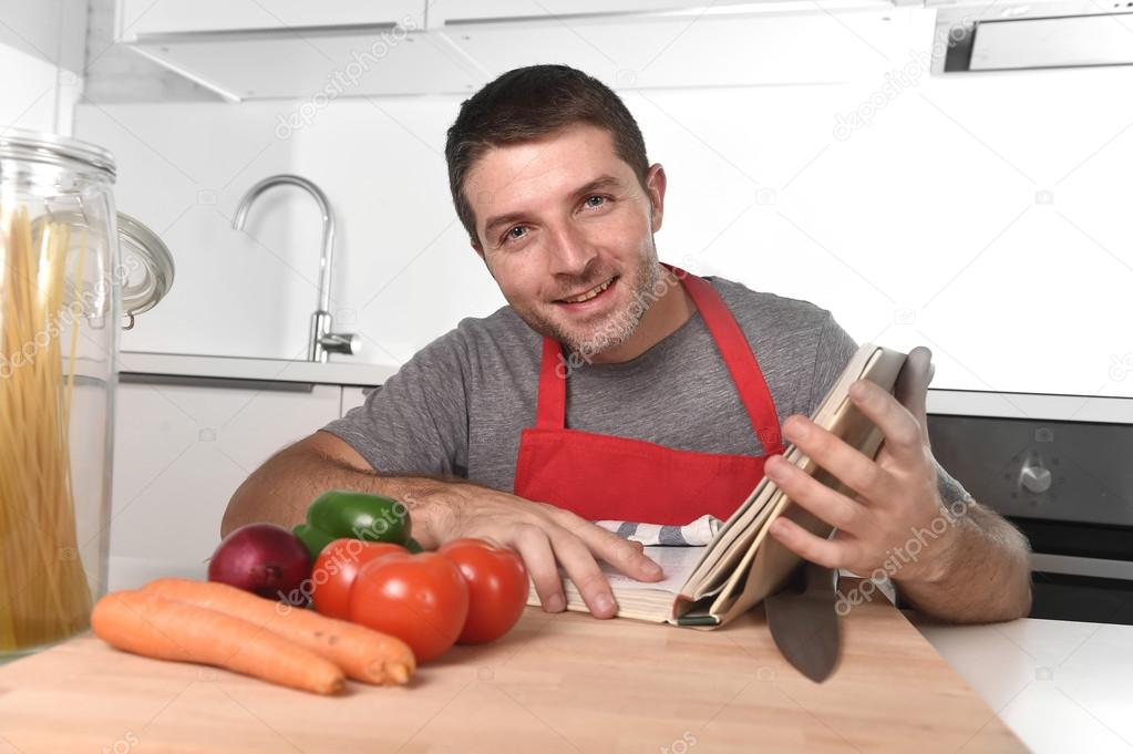 young happy man at kitchen reading recipe book in apron learning