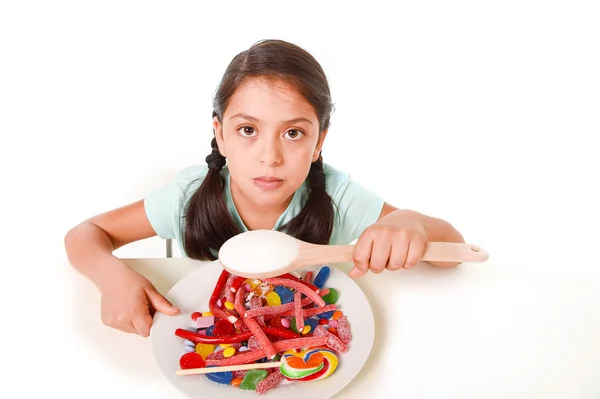Sad and vulnerable hispanic female child eating dish full of candy and gummies holding sugar spoon in wrong diet concept — 스톡 사진