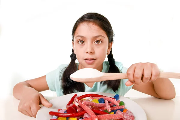 Triste e vulnerável hispânico fêmea criança comendo prato cheio de doces e gomas segurando colher de açúcar no conceito de dieta errada — Fotografia de Stock