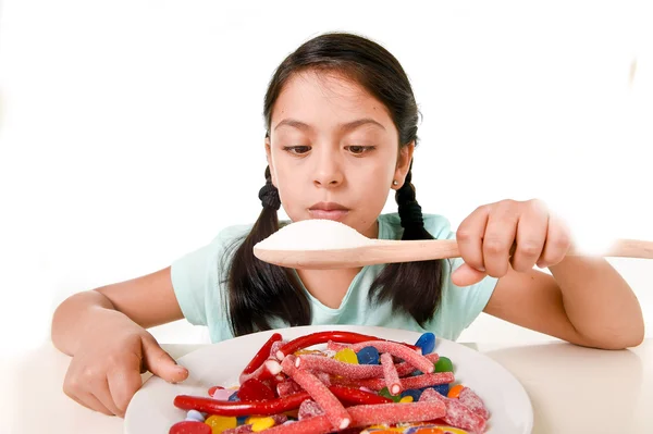 Triste e vulnerável hispânico fêmea criança comendo prato cheio de doces e gomas segurando colher de açúcar no conceito de dieta errada — Fotografia de Stock