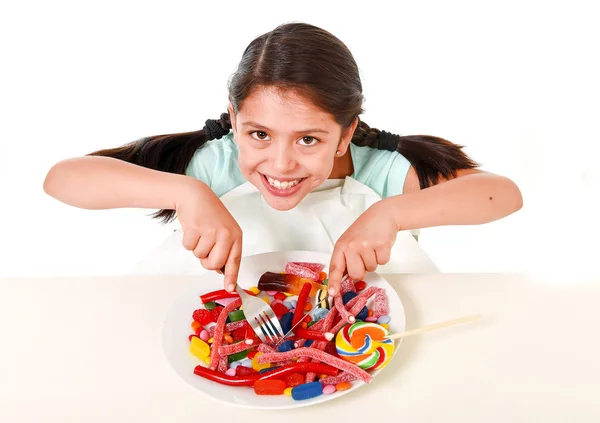 Happy Latin female child eating dish full of candy and gummies with fork and knife and big cola bottle in sugar abuse — Stock Photo, Image
