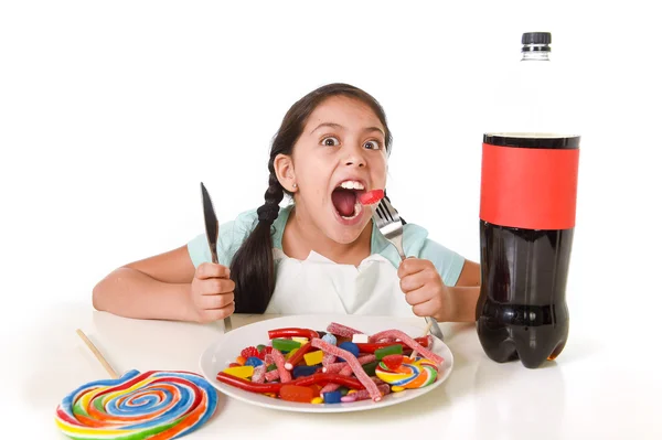 Criança latina feliz comendo prato cheio de doces e gomas com garfo e faca e garrafa de cola grande em abuso de açúcar — Fotografia de Stock