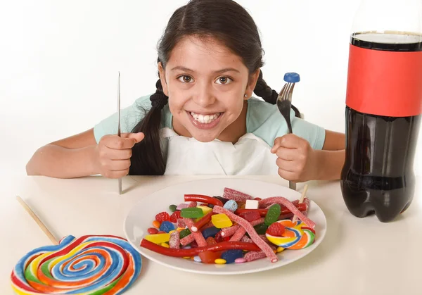 Счастливая латиноамериканская девочка ест блюдо, полное конфет и десен с вилкой и ножом и большой бутылкой колы в злоупотреблении сахаром — стоковое фото