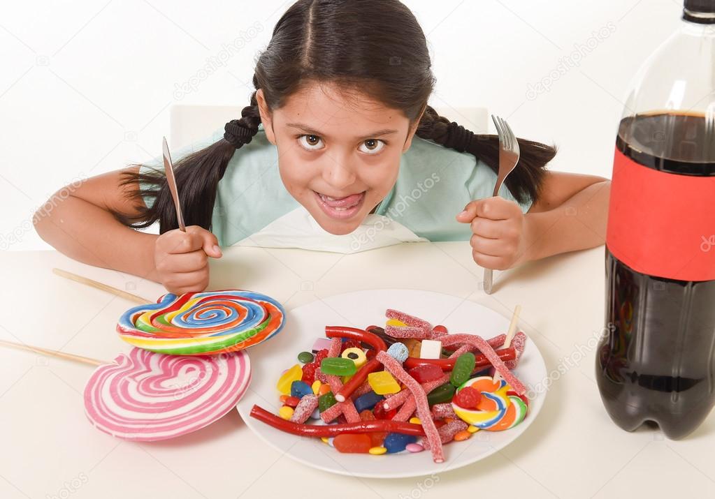 happy Latin female child eating dish full of candy and gummies with fork and knife and big cola bottle in sugar abuse