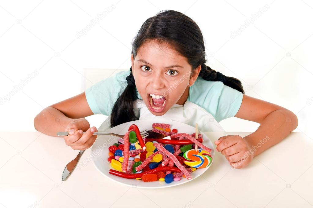 happy Latin female child eating dish full of candy and gummies w